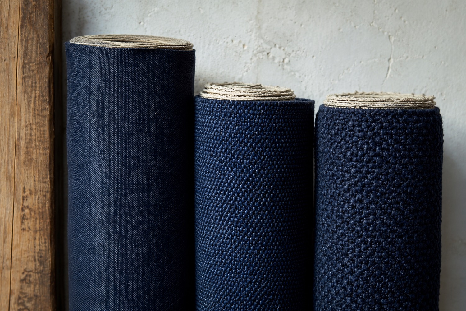 Three upright rolls of indigo cotton canvas of different weights standing against a pale plaster wall in daylight.