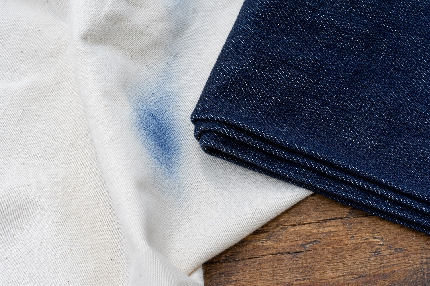 Folded raw indigo denim resting on pale unbleached cotton, with a faint blue smudge of transferred colour on the pale cloth.