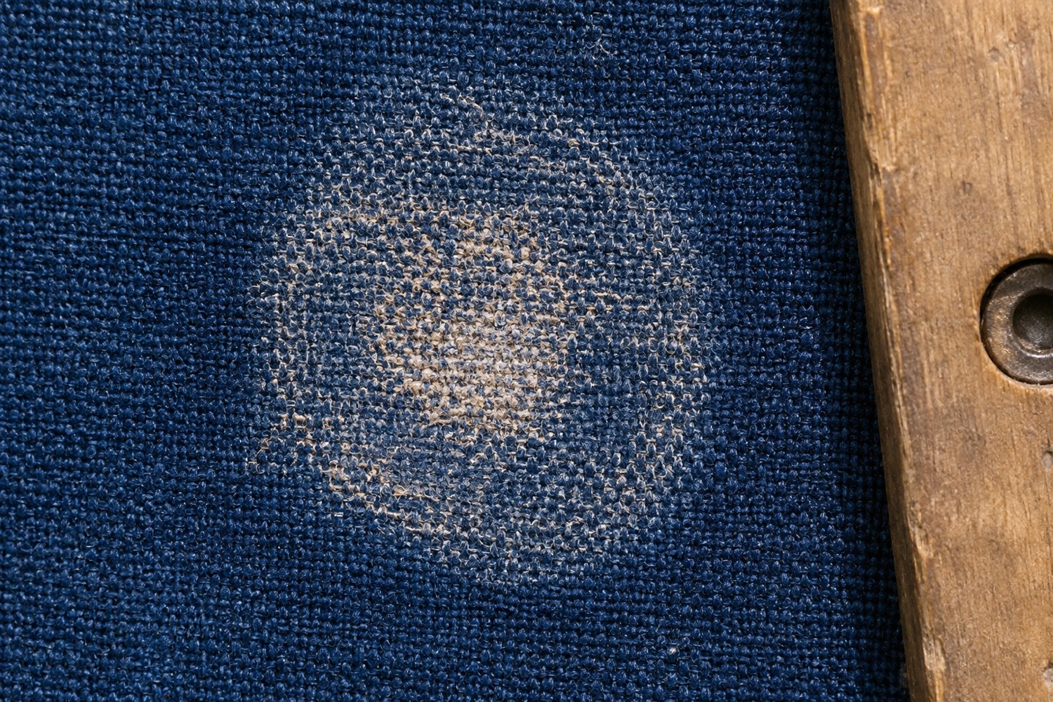 Macro view of a circular abrasion patch worn into indigo woven canvas, the worn circle showing lifted fibres against untouched weave.